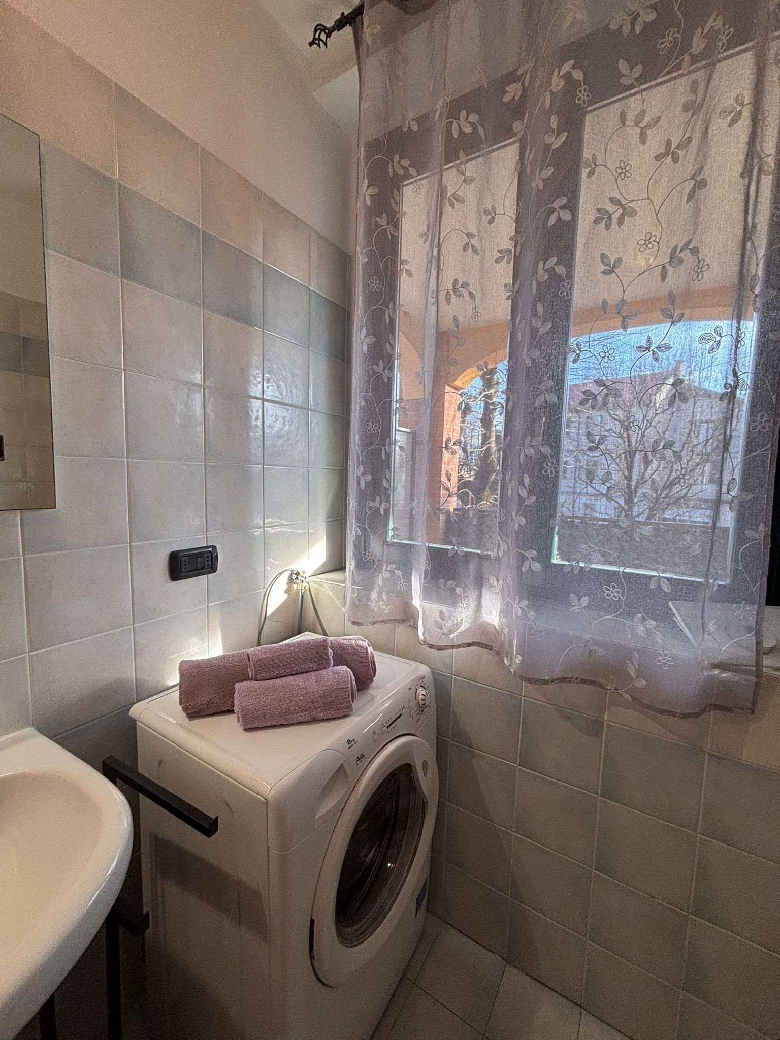 A compact bathroom with a washing machine, a sink, and a window with floral curtains.