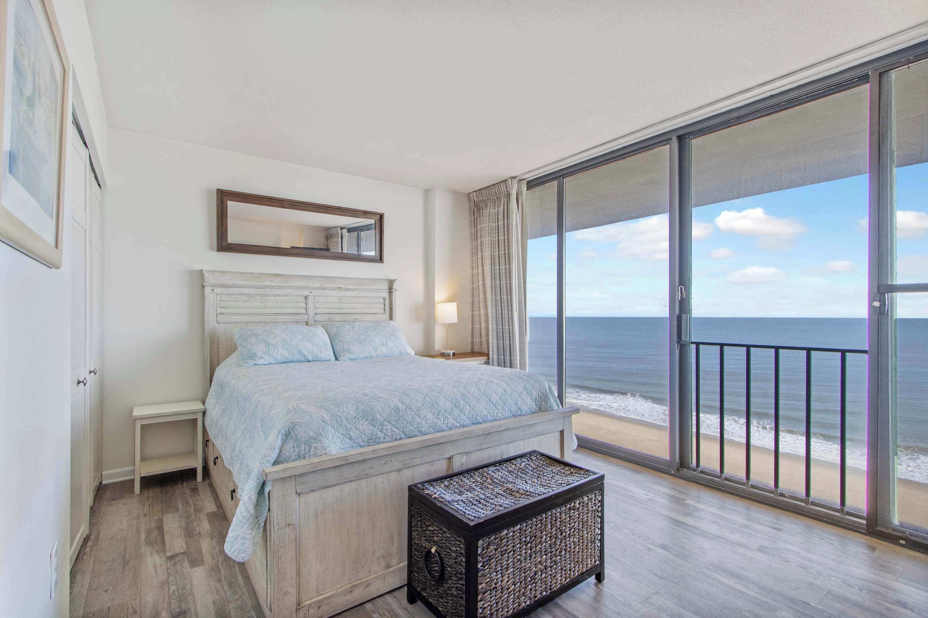 Ein Schlafzimmer mit einem Bett, einem Nachttisch, einem kleinen Hocker, einem großen Fenster mit Blick auf den Strand und einer Schiebetür.