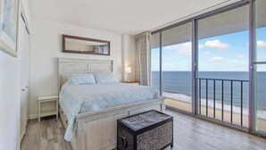 Ett sovrum med en säng, ett sängbord, en liten pall, ett stort fönster med utsikt över stranden och en skjutdörr i glas.