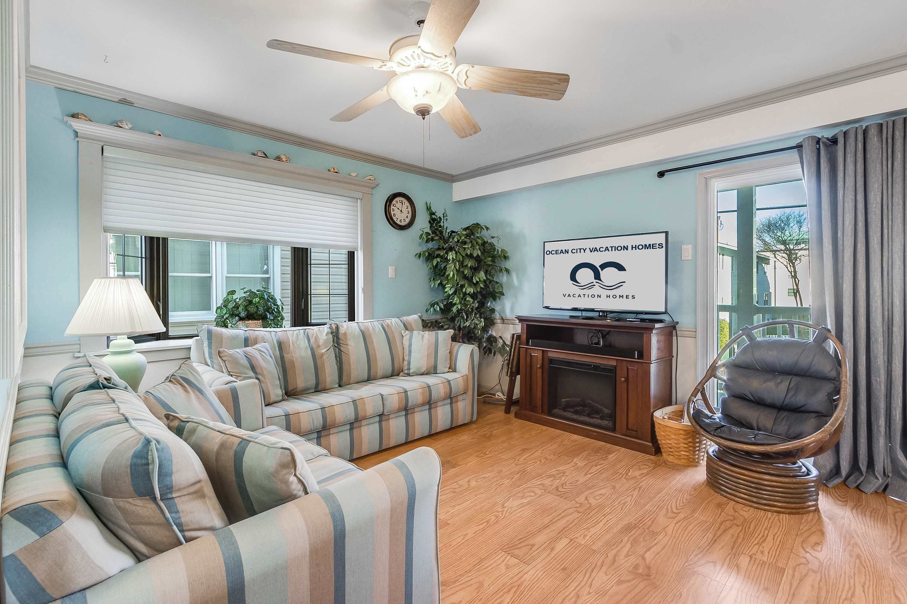 A living room with striped sofas, a wooden floor, a ceiling fan, a fireplace, and a sign for Ocean City Vacation Homes.