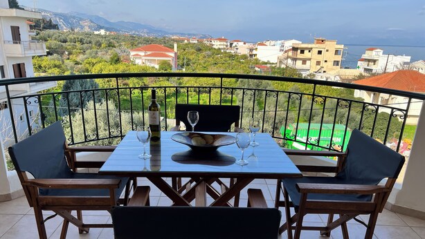 Un balcone con un tavolo apparecchiato per due, con bottiglia di vino e bicchieri. Il balcone si affaccia su una zona residenziale con case e vista sul mare.