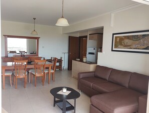 Ein Wohnzimmer mit braunem Sofa, eine Essecke mit Holztischen und -stühlen sowie eine Küchenzeile mit Kühlschrank und ein großes Fenster.