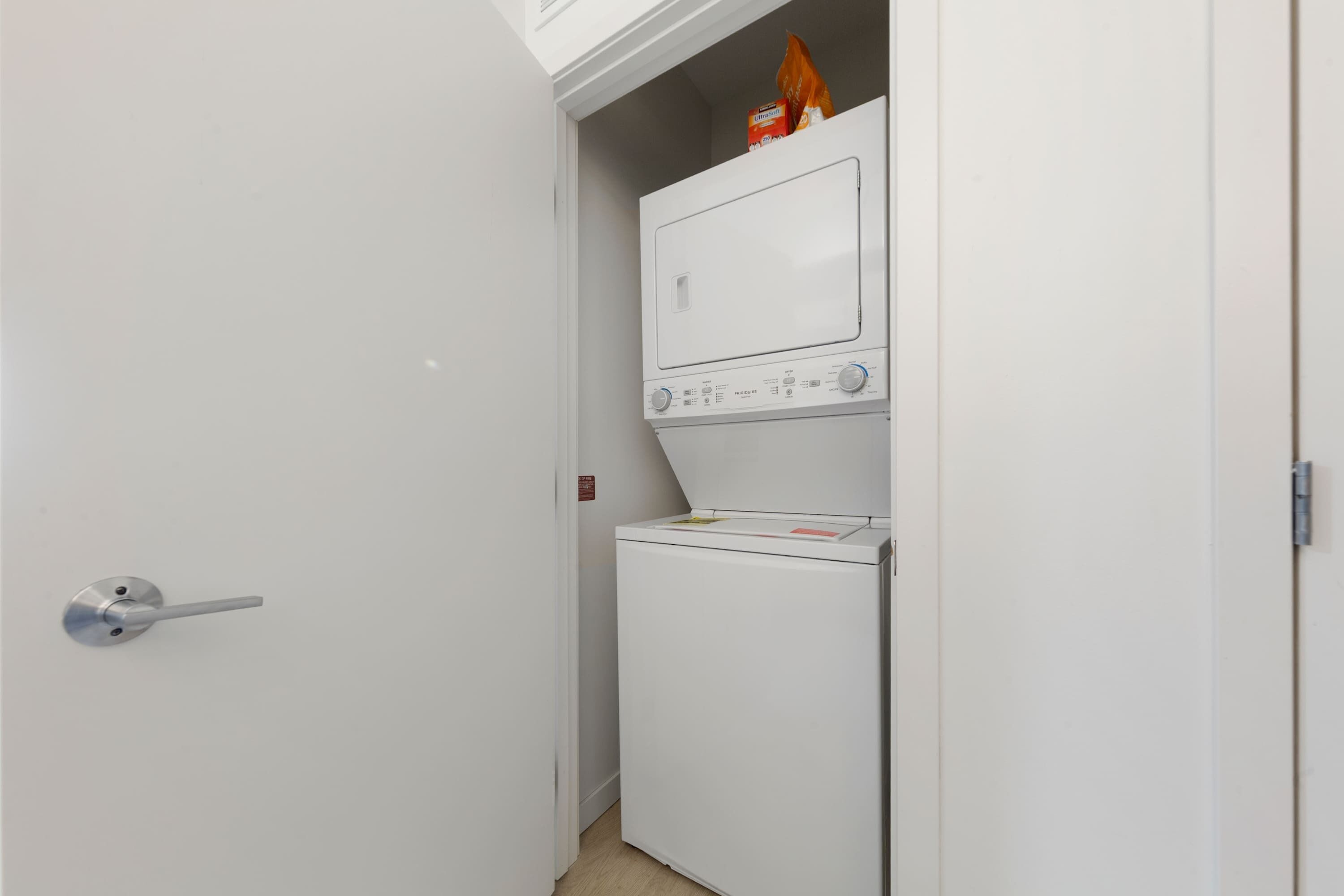 Une petite buanderie comprenant un lave-linge et un sèche-linge, une porte avec une poignée et une petite étagère sur laquelle se trouve un paquet de lessive.