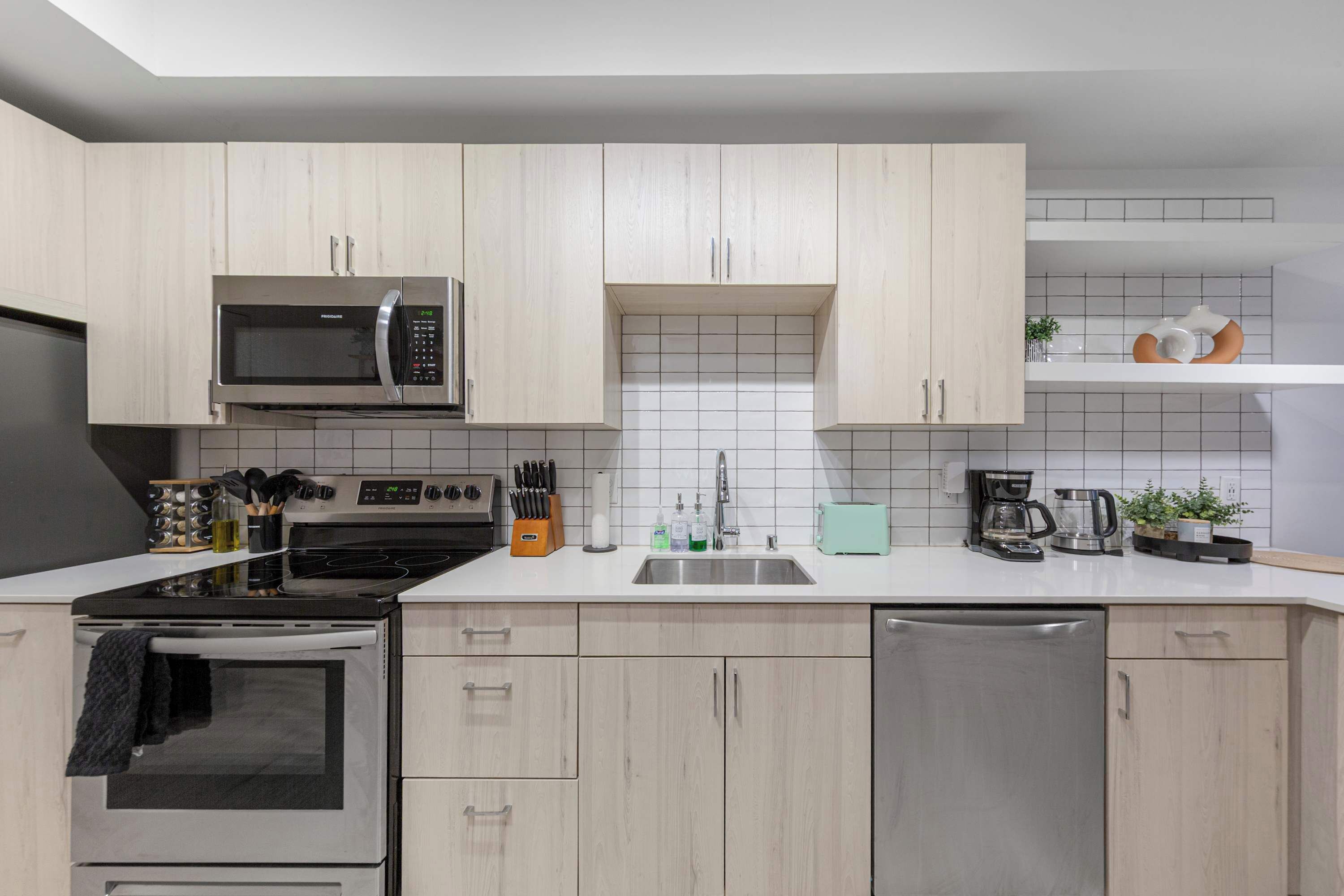 Une cuisine moderne avec des meubles en bois clair, des appareils électroménagers en acier inoxydable et un plan de travail blanc.