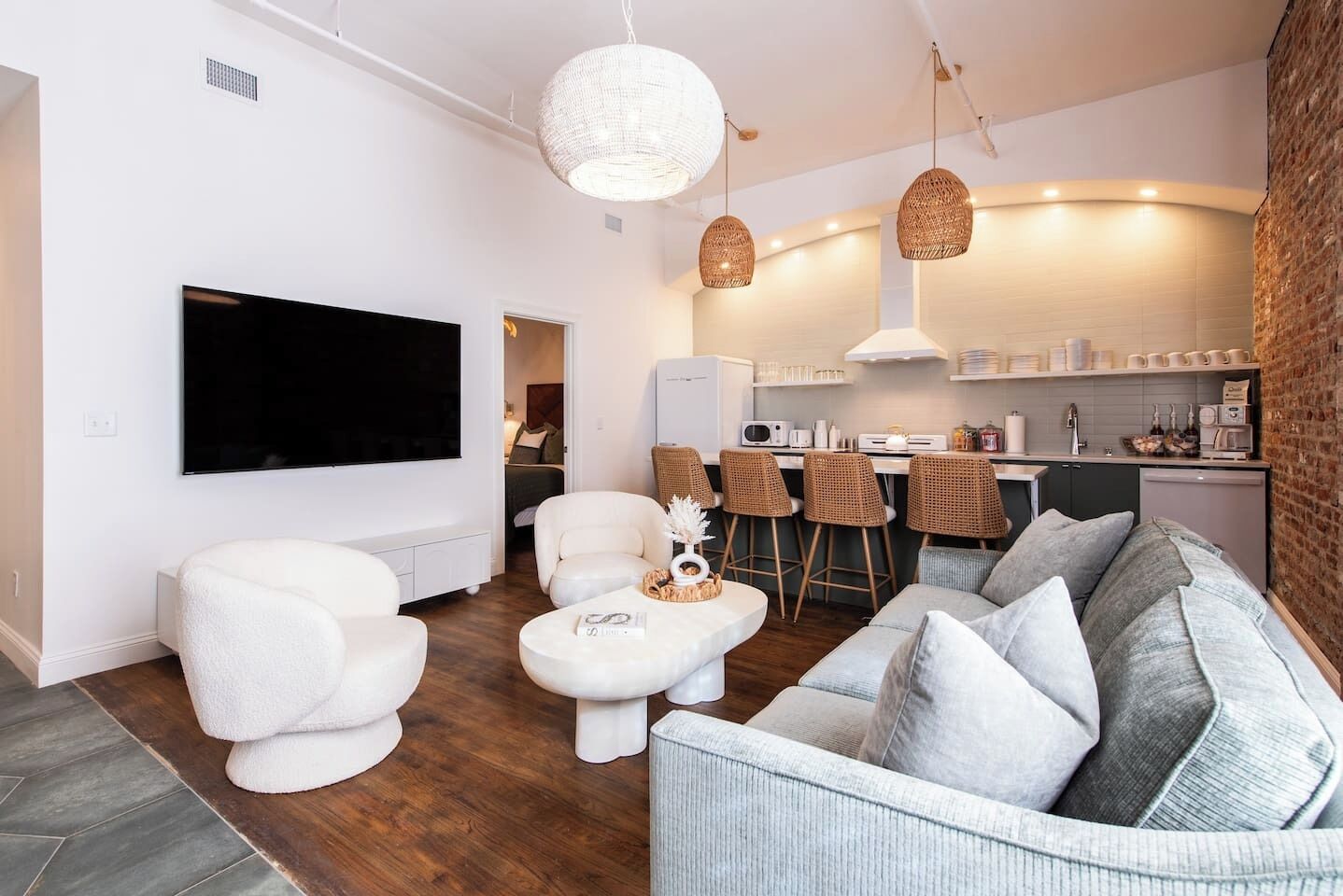 A modern living room with a flat-screen TV, white sofas, a coffee table, wicker pendant lights, and a kitchen area with bar stools.
