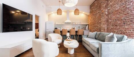 Una sala de estar moderna con televisor de pantalla plana, muebles blancos, pared de ladrillo y una zona de cocina.