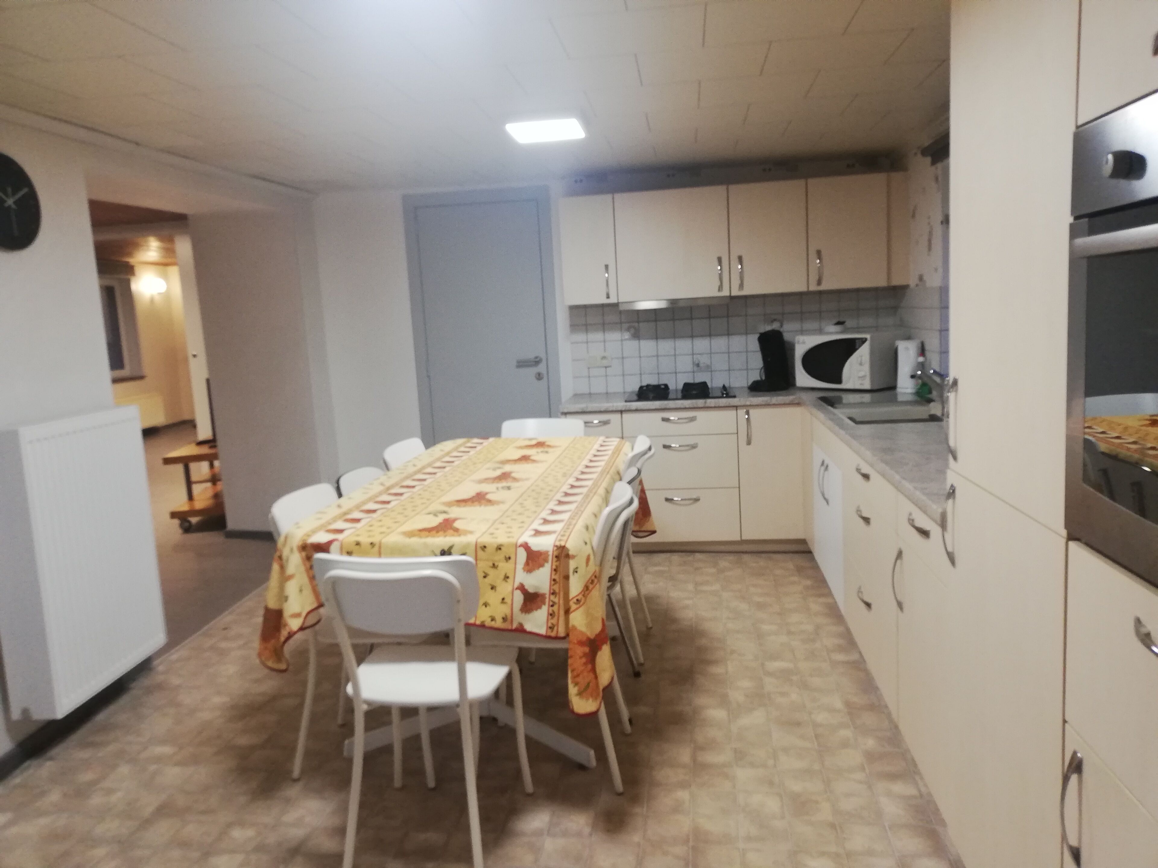 Een kleine, praktische keuken met witte kastjes, een magnetron en een fornuis. Er staat een eettafel met stoelen en een tafelkleed met een patroon.