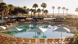 Zona de piscina con sillones de descanso y sombrillas, rodeada de palmeras y vista al océano.