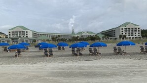 Un hotel con varios pisos y una torre central, con numerosas sombrillas y sillas de playa azules.