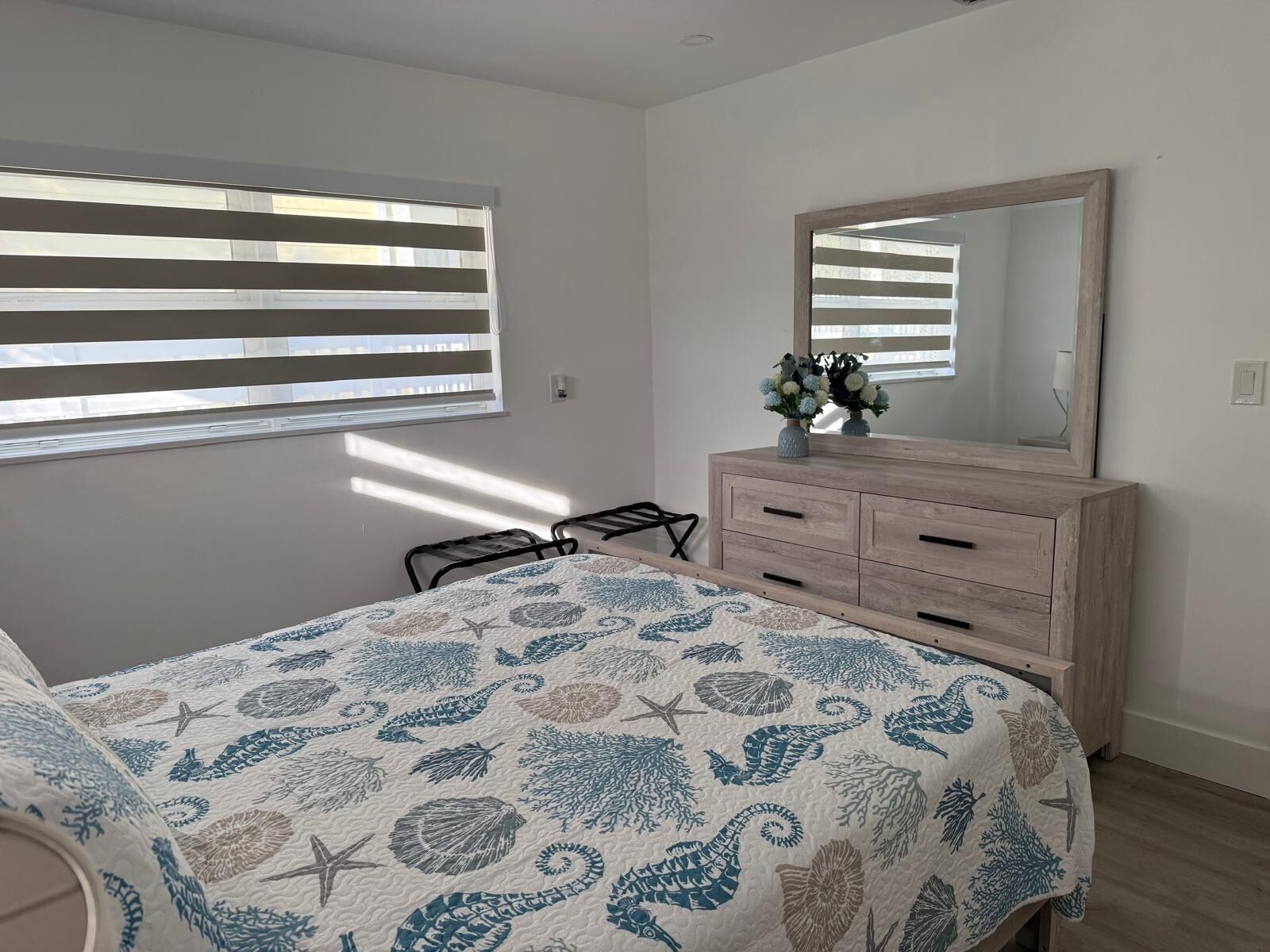 Ein Schlafzimmer mit Bett, Kommode, Spiegel und einem Fenster mit Jalousien.