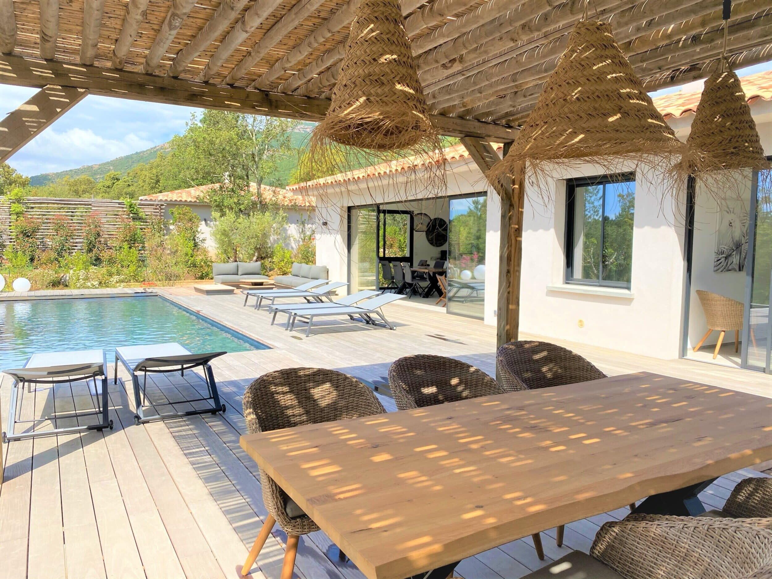 A poolside area with a wooden deck, lounge chairs, a table, and a pergola with thatched umbrellas.