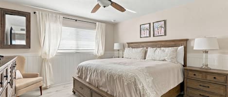 A bedroom with a bed, bedside tables, a dresser, a ceiling fan, a window with curtains, and artwork on the wall.