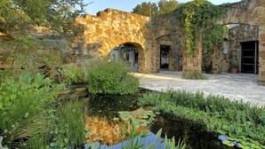 Un patio al aire libre con un arco de piedra, un pequeño estanque con nenúfares y abundante vegetación circundante.