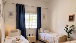 Quarto com duas camas, uma janela com cortinas, uma mesa de cabeceira com abajur e um ar condicionado fixado na parede.