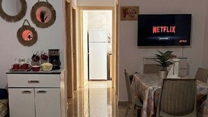 Uma sala de estar com uma TV exibindo 'NETFLIX', uma área de jantar com mesa e cadeiras, uma cozinha com armários e cafeteira, e uma pequena mesa redonda com controle remoto e vela.