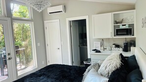 A modern bedroom with a large window, a black furry rug, a bed with white and black pillows, and a kitchenette with a microwave and coffee maker.