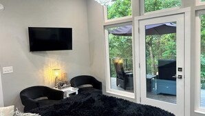 Una habitación con televisión de pantalla plana, una lámpara, dos sillas, una mesita y un ventanal con vistas a la vegetación.
