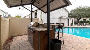 Un espace barbecue extérieur équipé d’un gril, d’un coin salon abrité et d’une piscine.
