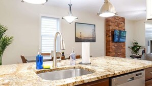 Une cuisine dotée de plans de travail en granit, d’un évier en acier inoxydable et d’un lave-vaisselle. On y trouve un mur décoratif en briques, un tableau représentant le Golden Gate Bridge et un luminaire suspendu.