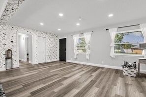 A spacious room with a wooden floor, white walls, and a geometric patterned wallpaper. There are two windows with white curtains, a door, and a small table with a lamp. - Resort Like Winter Park Pool Home (Winter Park)