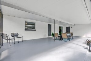 Een ruime binnenruimte met een witte bakstenen muur, een raam en een deur. Er staan metalen stoelen en een tafel met een plantje.