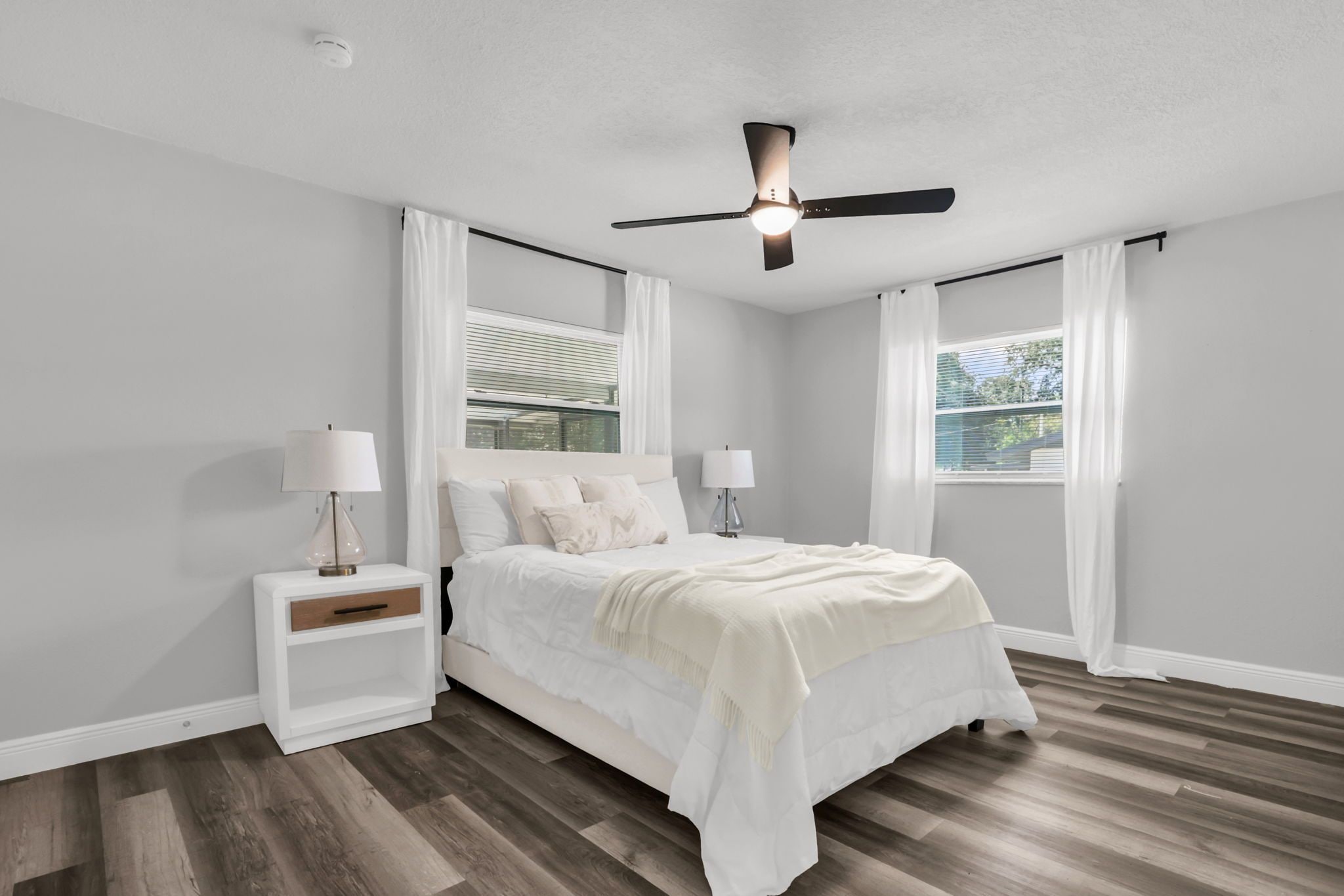 A bedroom with a bed, nightstands, a ceiling fan, and windows with curtains.