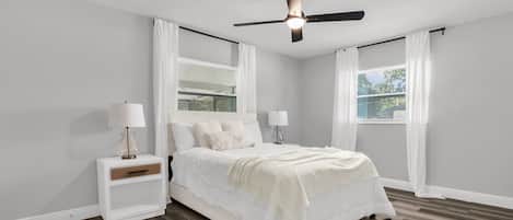 Un dormitorio con cama, mesitas de noche, ventilador de techo y ventanas con cortinas.