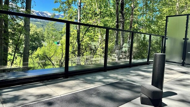 Un espace extérieur moderne doté d’une balustrade en verre, d’un banc en pierre et offrant une vue sur une forêt et des montagnes.
