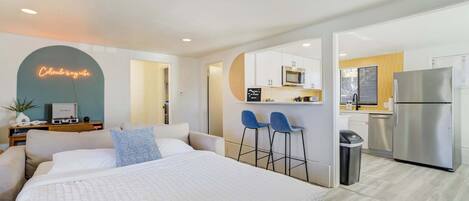 A modern hotel room with a large bed, a neon sign, a kitchen area with a refrigerator, and a bar stool.