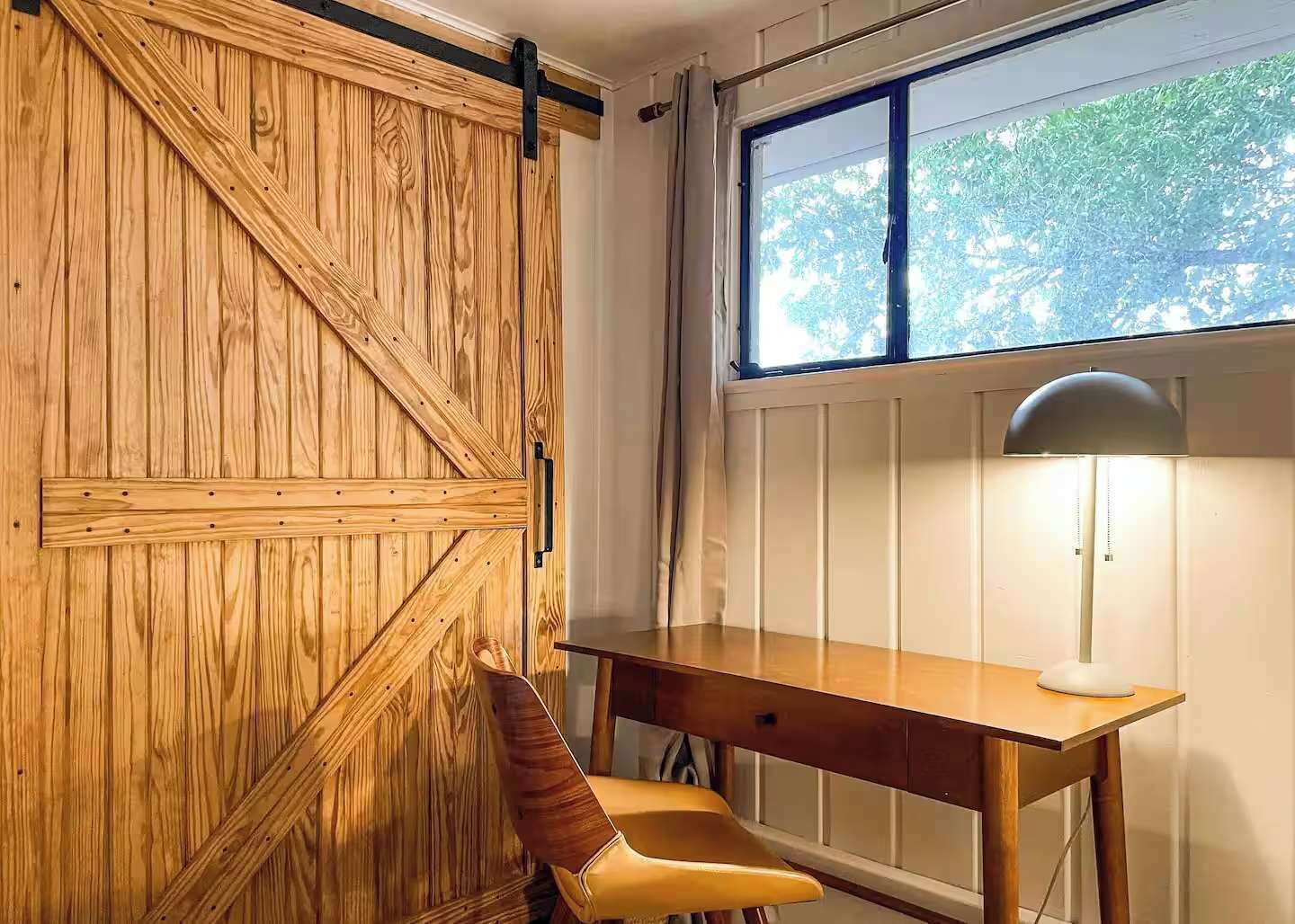 Une pièce avec un mur lambrissé en bois, un bureau avec une chaise, une fenêtre donnant sur des arbres et un radiateur.