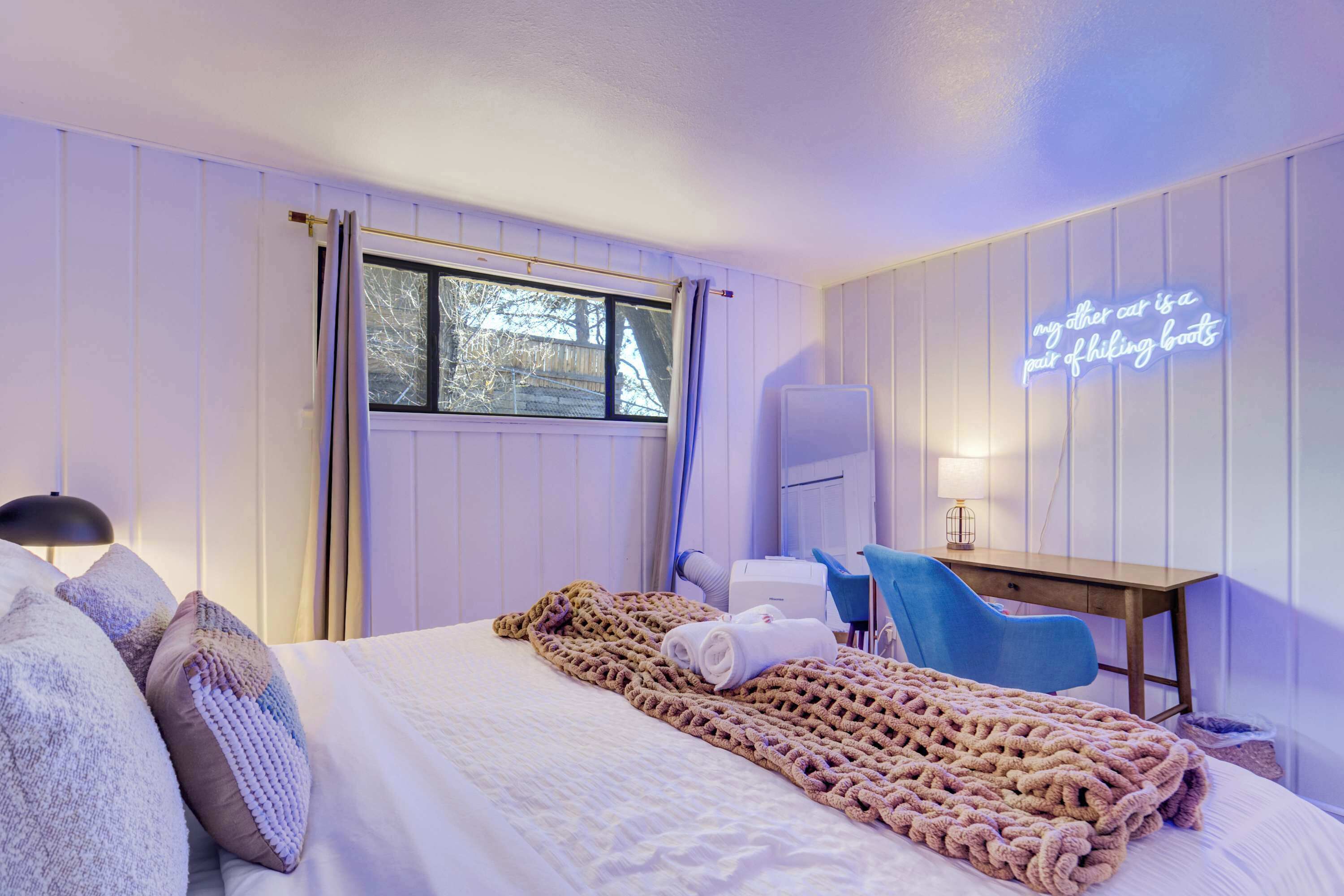 Une chambre d’hôtel avec un lit, un bureau, une chaise, une fenêtre et un néon sur le mur.