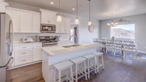 A modern kitchen with white cabinetry, a central island with bar stools, and a dining area with white chairs. - Premium Family Escape: Heated Pool, Private Hot Tub, Fire Pit & Games (Hurricane)