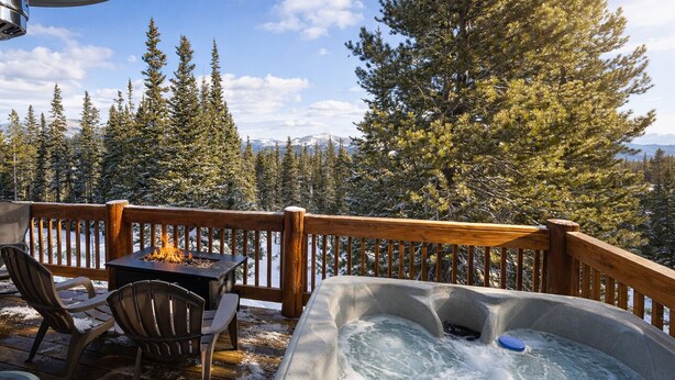 Une terrasse en bois avec un jacuzzi, un foyer et des chaises, entourée d’arbres couverts de neige et avec des montagnes en arrière-plan.