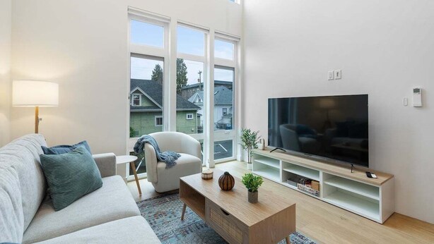 Ein modernes Wohnzimmer mit einer Couch, einem Couchtisch, einem Fernseher und einem Fenster mit Blick auf Häuser.