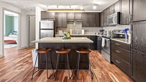 Una cocina moderna con armarios de madera oscura, electrodomésticos de acero inoxidable, una isla central con taburetes y una lámpara colgante que proporciona luz.