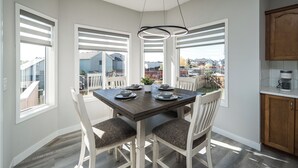Comedor con una mesa preparada para cuatro, sillas blancas, lámpara colgante y ventanas amplias con persianas.