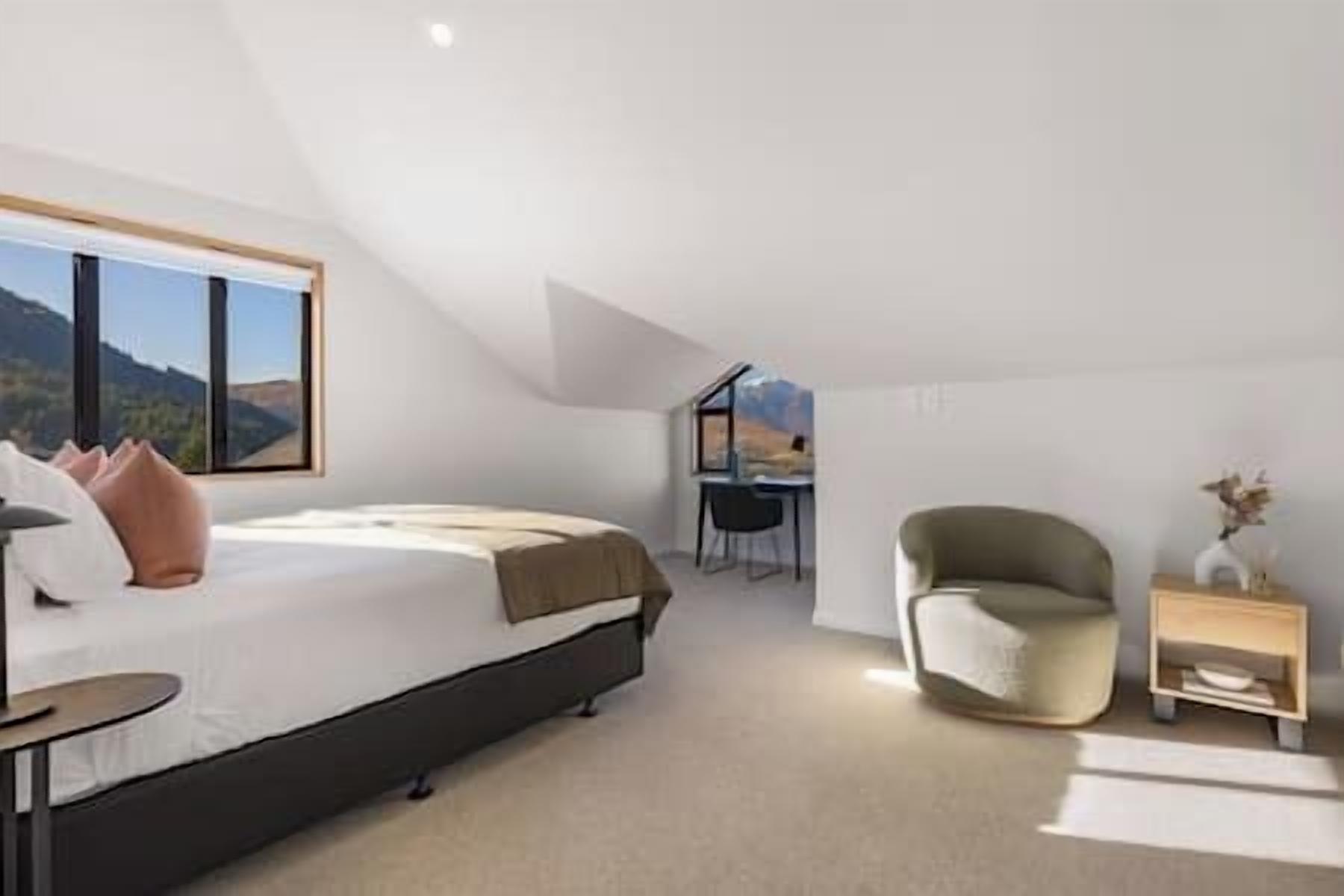 A modern bedroom with a large bed, a desk, and a chair, featuring a view of mountains through large windows.