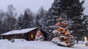 Eine Blockhütte mit schneebedecktem Dach, umgeben von immergrünen Bäumen und einem beleuchteten Weihnachtsbaum.
