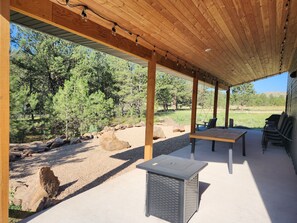 An outdoor covered patio with a table and chairs, surrounded by trees and a gravel path.