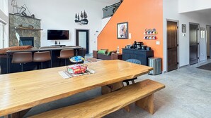 A modern kitchen with a stone fireplace, a dining area with a wooden table and benches, and a kitchen island with a coffee machine. - New Black Hills Home Next to Wind Cave National Park (Custer)