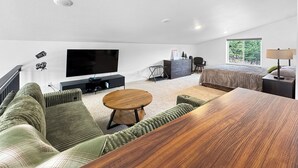A modern living room with a flat-screen TV, a sofa, a coffee table, a bed, and a window with a view of trees. - New Black Hills Home Next to Wind Cave National Park (Custer)