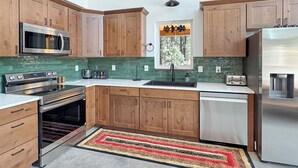 A kitchen with wooden cabinets, stainless steel appliances, a green backsplash, and a window with a view of trees. - New Black Hills Home Next to Wind Cave National Park (Custer)
