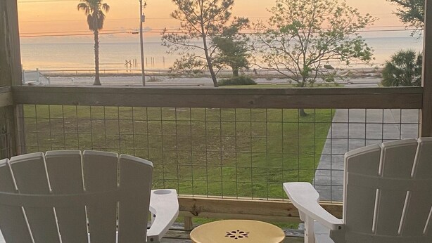 A wooden deck with two white chairs and a yellow table, overlooking a beach with trees and a sunset.