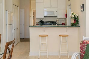 Une cuisine de taille réduite avec des meubles blancs, un four à micro-ondes, une cuisinière et deux tabourets en bois.