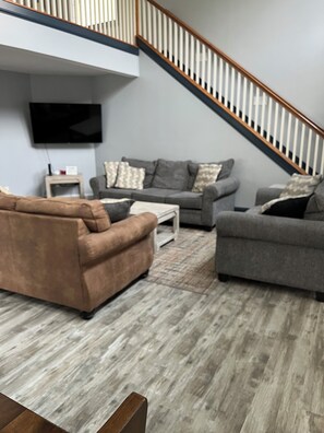 A modern living room with a grey sofa set, a brown sofa, and a wooden coffee table. A staircase with a wooden railing is visible in the background.