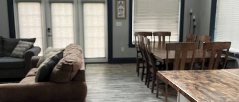 A spacious living area with a sofa, dining table, and chairs. There are large windows with blinds and a door with a window.