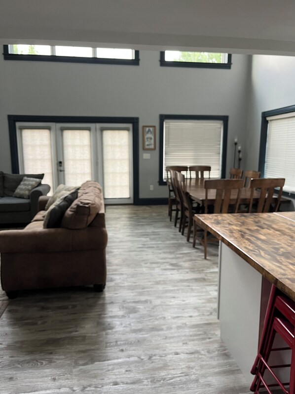 A spacious living area with a sofa, dining table, and chairs. There are large windows with blinds and a door with a window.