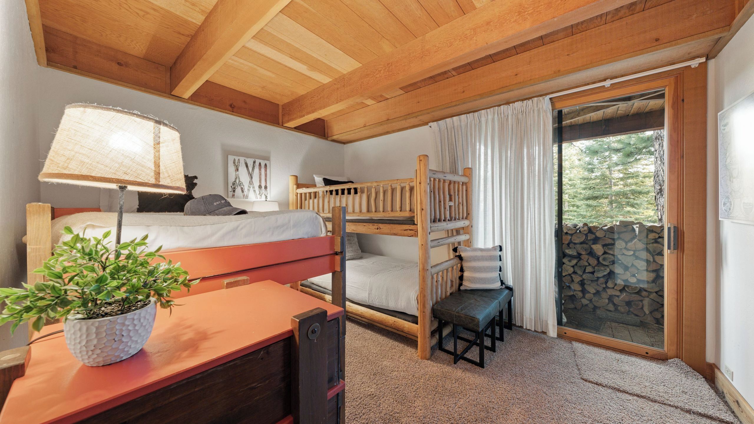 A room with a bunk bed, a desk with a lamp, a potted plant, and a view of a stone wall and trees through a glass door.