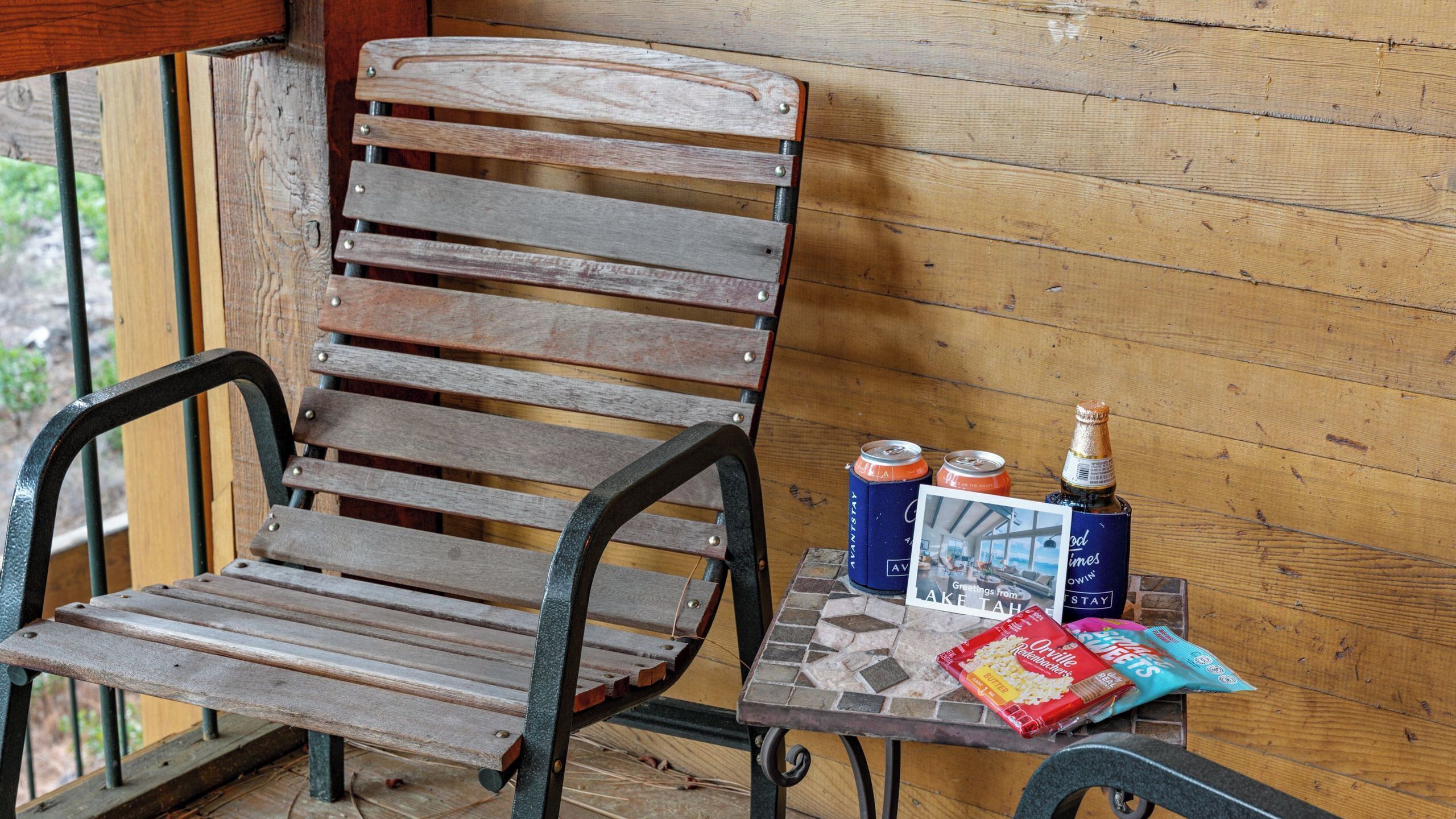 Una silla y una mesa de madera con una botella, latas y una postal en un porche.