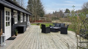 Une terrasse en bois avec des meubles d’extérieur, une maison aux boiseries blanches et une vue sur le paysage environnant.